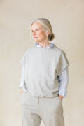 Woman wearing a light gray sweater over a blue shirt with beige pants against a plain background