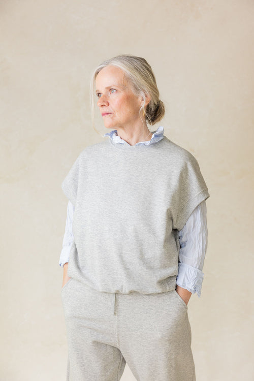 Woman wearing a light gray sweater over a blue shirt with beige pants against a plain background