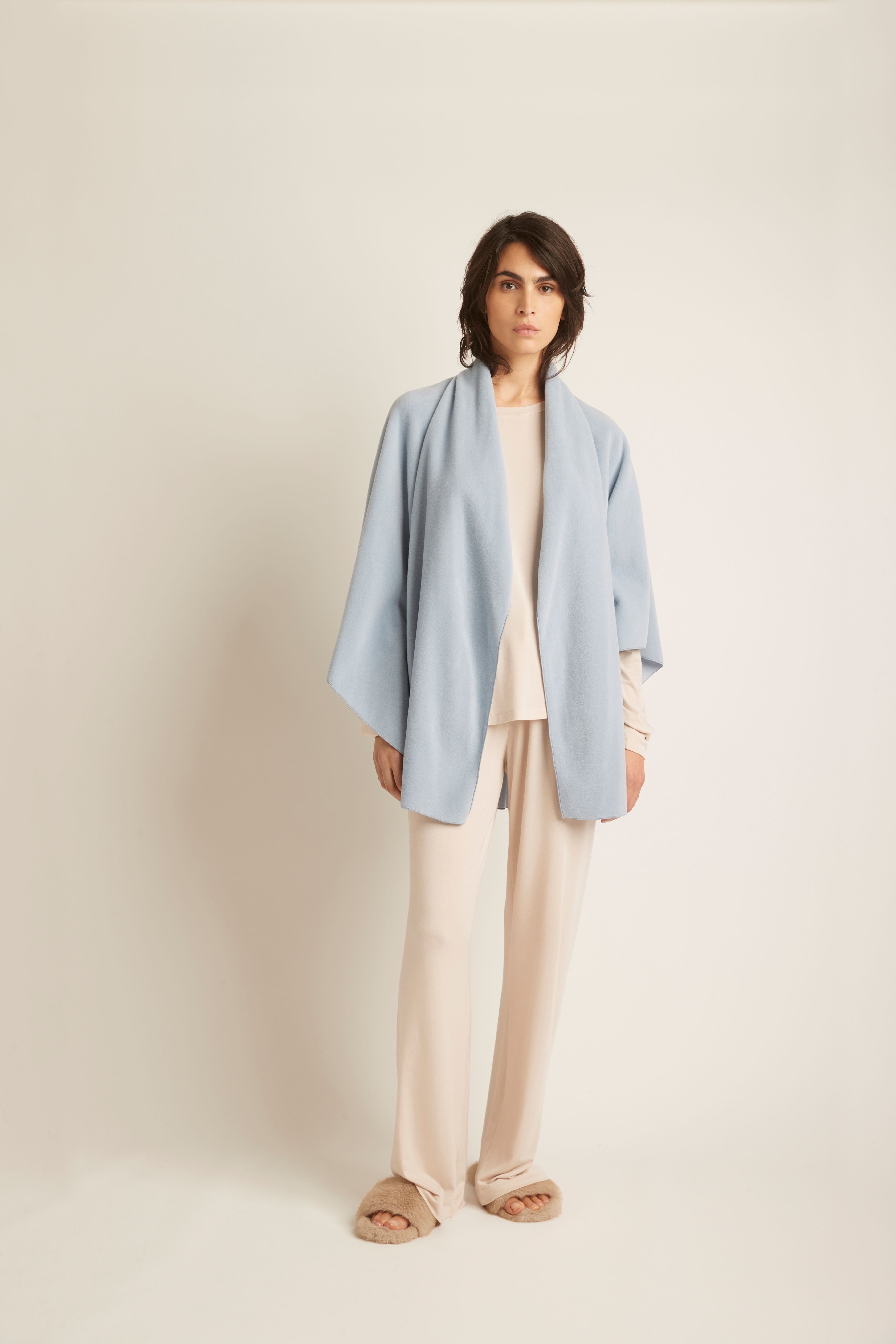 A woman stands against a plain warm off-white studio background wearing a light blue open-front draped jacket over a cream top and matching cream wide-leg pants, with tan slippers.