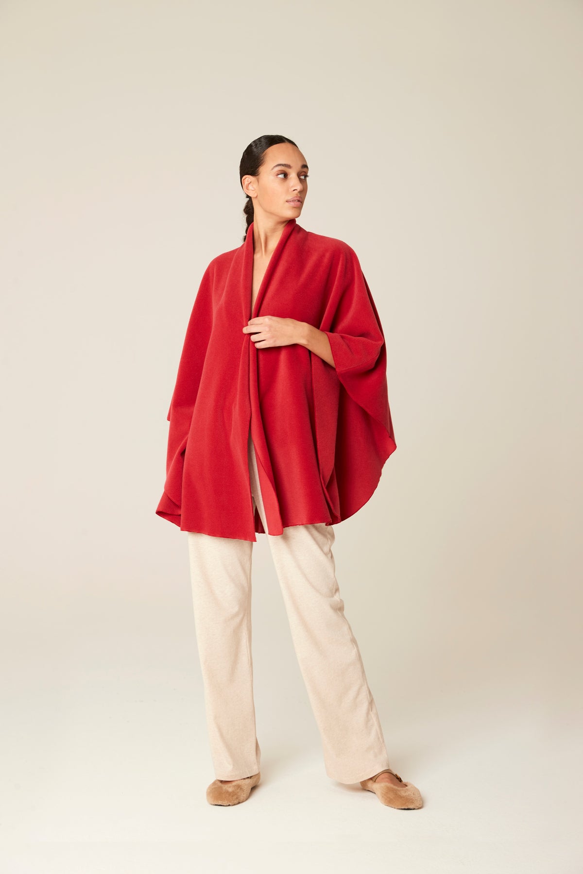 A woman modeling a vibrant red draped wrap jacket or poncho over cream-colored wide-leg trousers and tan shoes, set against a plain light beige studio background.
