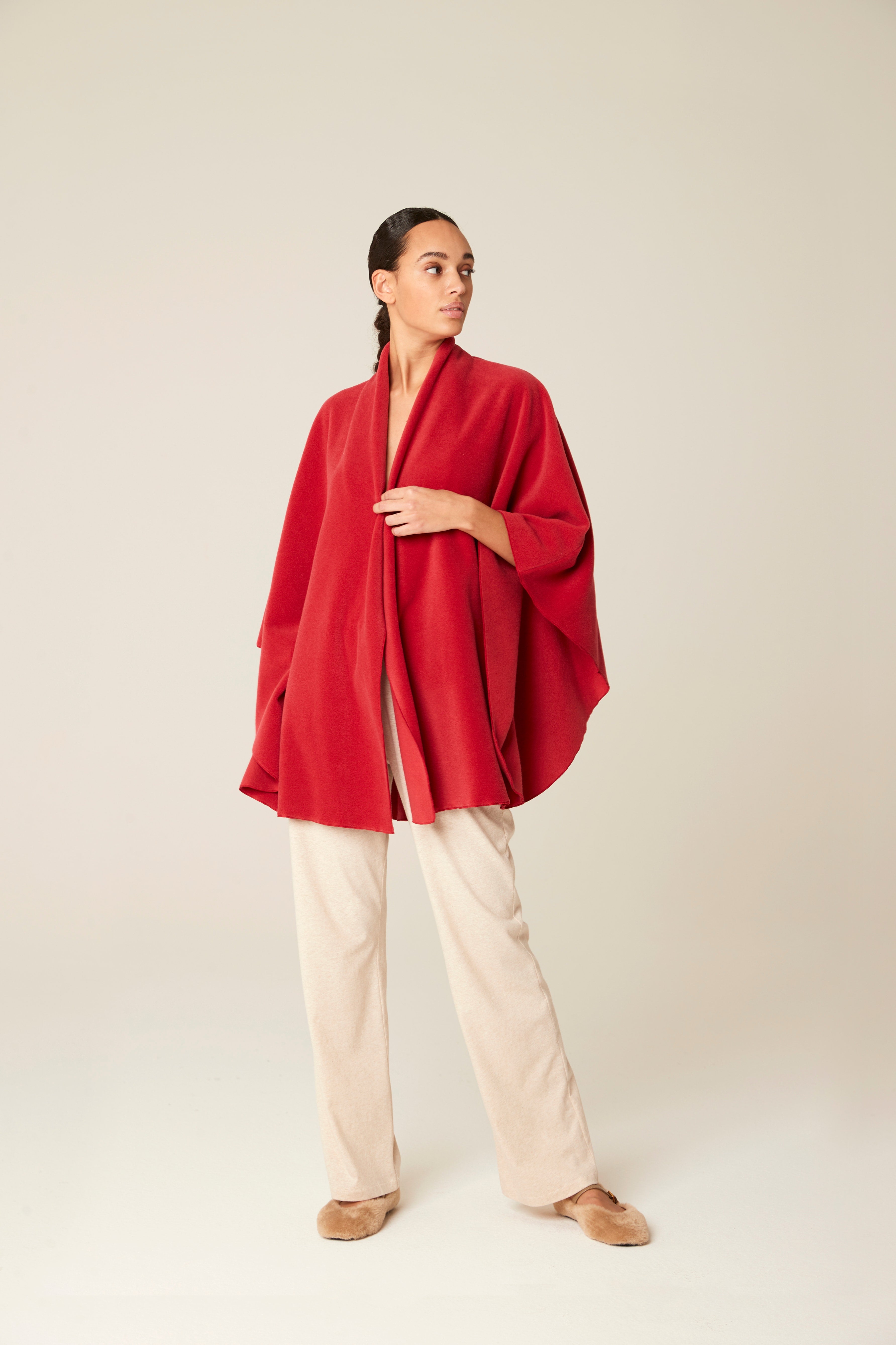 A woman modeling a vibrant red draped wrap jacket or poncho over cream-colored wide-leg trousers and tan shoes, set against a plain light beige studio background.