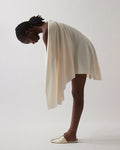 A model wearing a draped, asymmetrical cream-colored wrap dress or shawl with matching slip-on shoes, captured in a side-profile pose against a neutral light gray studio background.