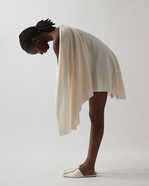 A model wearing a draped, asymmetrical cream-colored wrap dress or shawl with matching slip-on shoes, captured in a side-profile pose against a neutral light gray studio background.