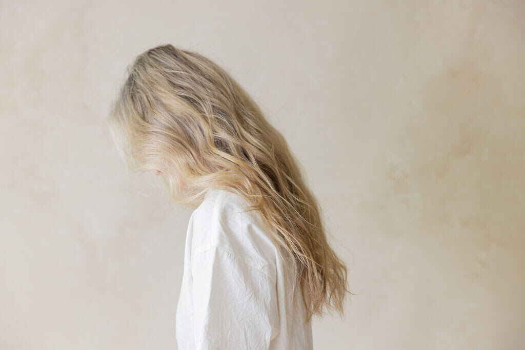 Woman with long blond hair, facing sideways against a cream wall wearing a white shirt. 